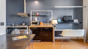 A compact kitchen with a wooden countertop, a built-in stove, and a microwave. There is a dining table with chairs and a white bench. A TV is mounted on the wall, and a shelf with various items is above the counter. - Piazza Santa Croce Design Apartment with Balcony (Florence)