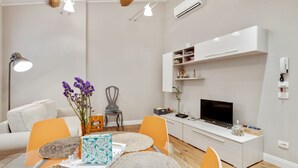 A modern living room with a glass dining table, orange chairs, a white sofa, and a TV on a white cabinet.