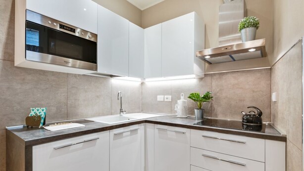 A modern kitchen with white cabinetry, a stainless steel oven, a sink, and a countertop with a kettle and potted plants.