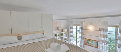 A bedroom with a large bed, a wardrobe, a desk, and a view of greenery through the window.