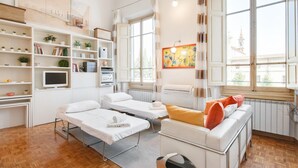 A clean, well-lit room with two beds, a sofa, a desk, and shelves with plants and books. - Piazza San Marco Studio Flat (Florence)