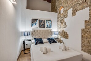 A bedroom with a tufted headboard, a bed with white linens, two blue patterned pillows, a nightstand with a lamp, and three framed artworks above the bed.