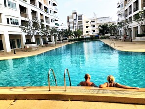A swimming pool surrounded by multi-story buildings with balconies, a clear sky, and a few people relaxing by the pool. - Platinum Suites Studio Condo Jomtien - Sleeps 2 (Pattaya)