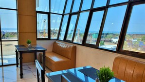 A modern room with a glass table, leather sofa, and large windows offering a view of the sea and parked cars. - VITS Excellensea Karde Beach (Dapoli)