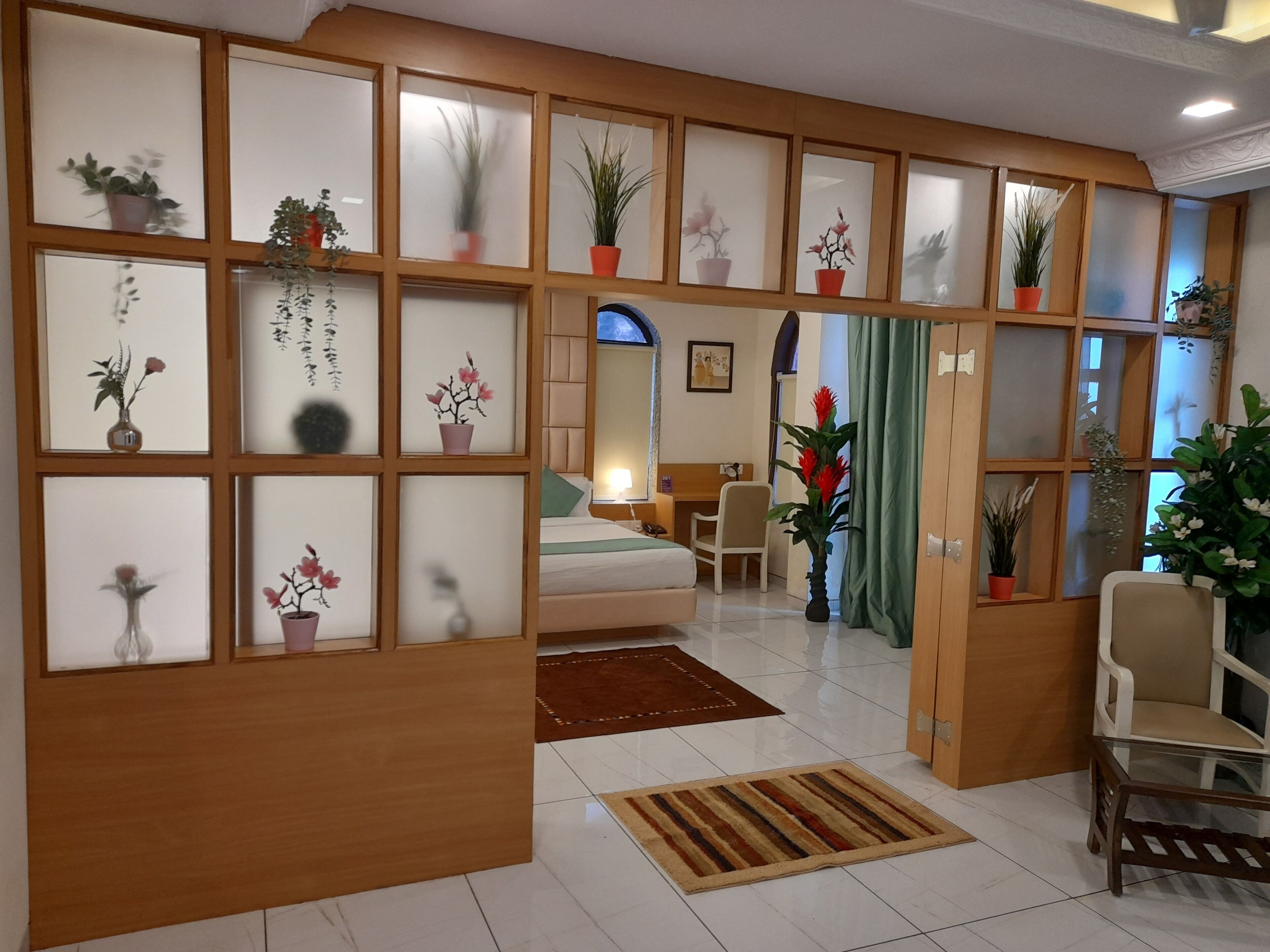 A hotel room with a wooden partition, a bed, a desk, a chair, and various potted plants.