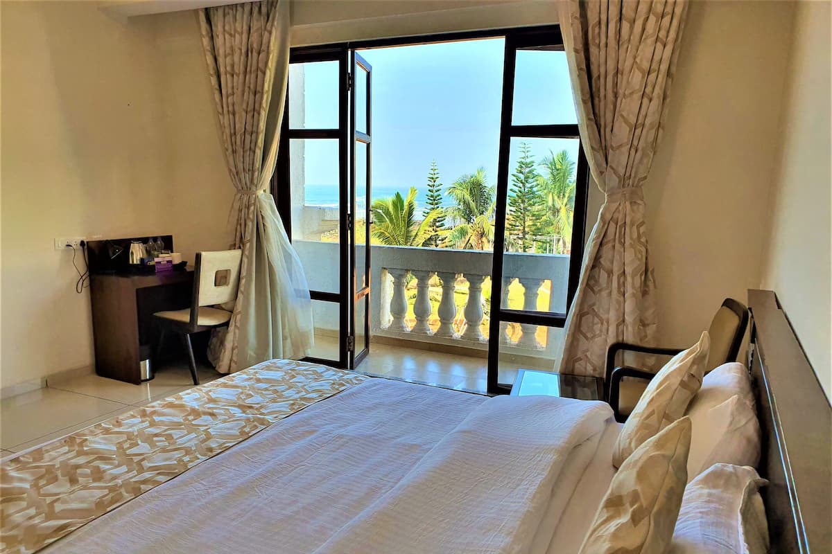 A hotel room with a bed, a desk, and a chair. There is a balcony with a view of the ocean and palm trees.
