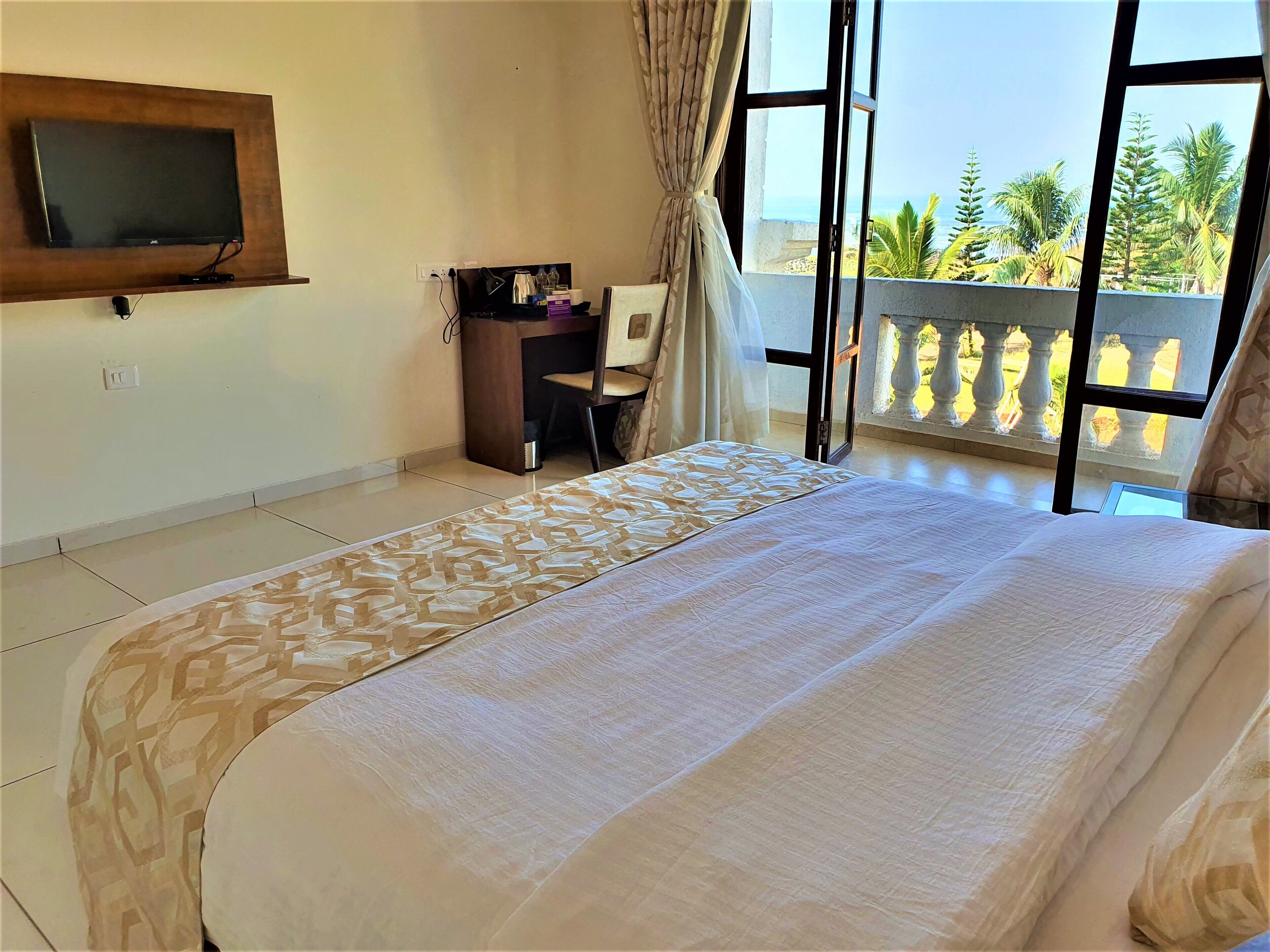 Habitación de hotel con cama, escritorio con silla, televisor en la pared y balcón con vistas a palmeras y al mar.