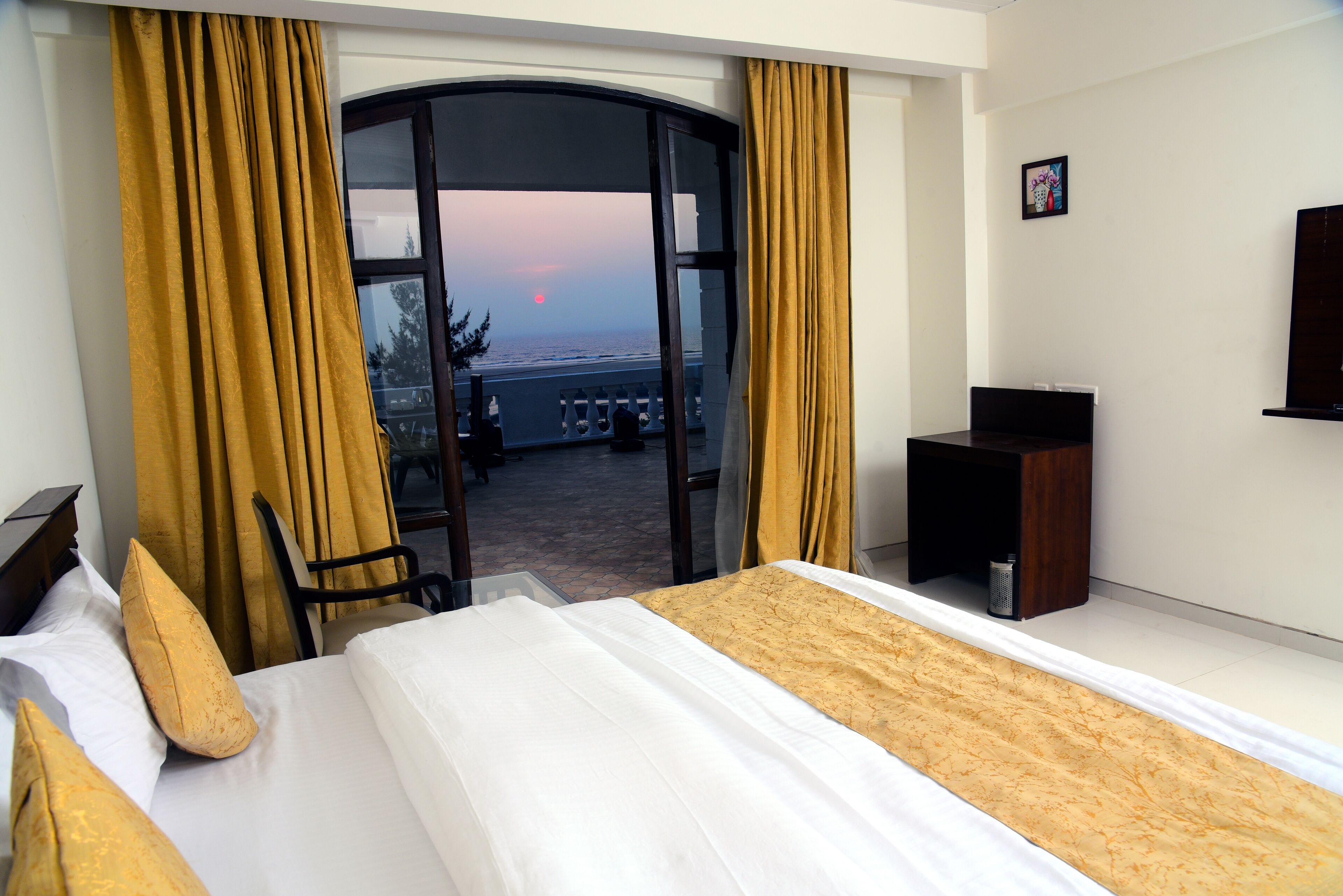 A hotel room with a bed, a desk, a chair, and a view of the ocean at sunset.