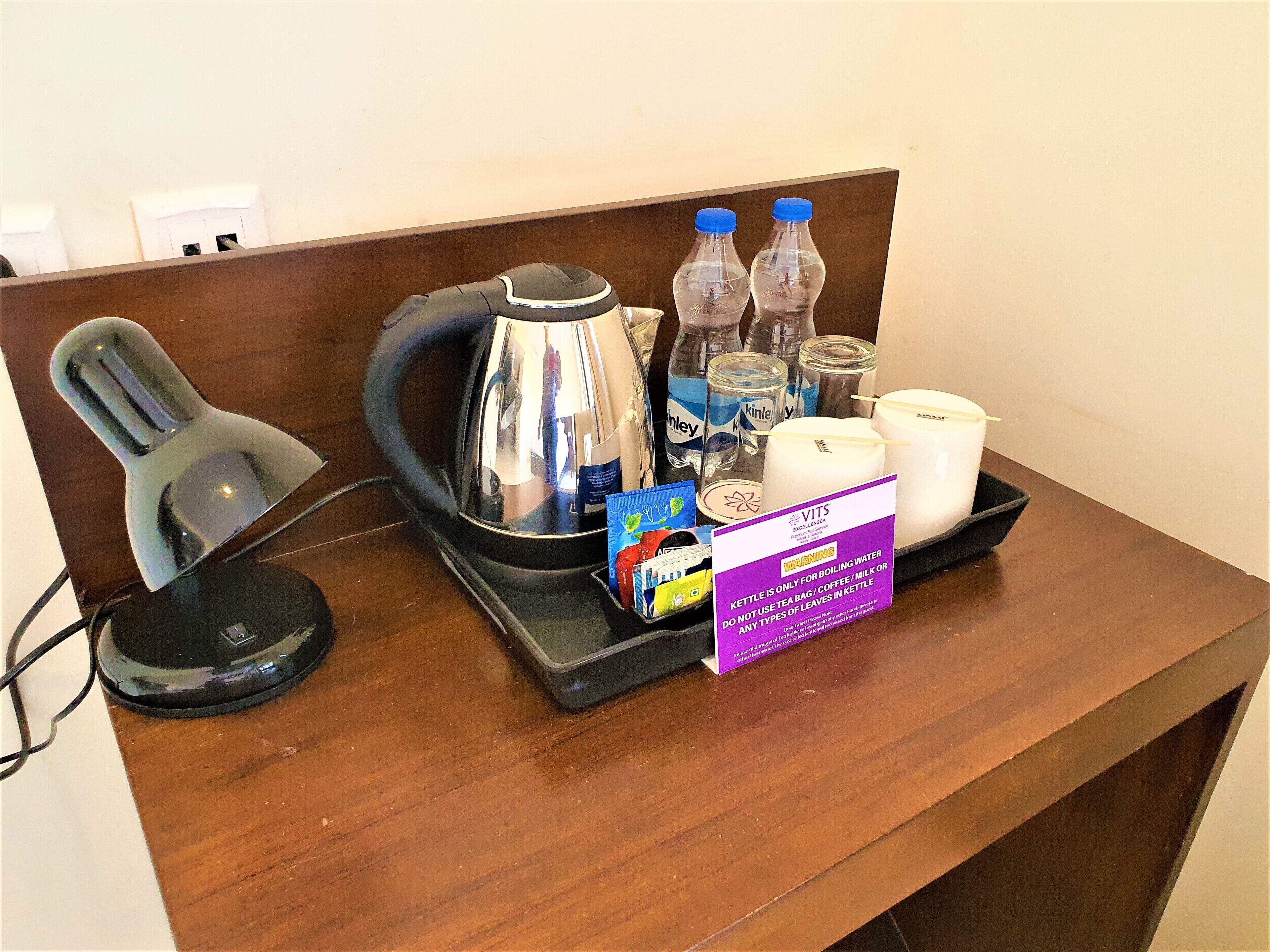 Un comptoir d’accueil d’hôtel avec une bouilloire, des bouteilles d’eau, une bougie, une boîte de sachets de thé et un panneau indiquant que la bouilloire est réservée à la préparation de café ou de thé.