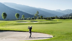 A golf course with a player in a sand bunker, surrounded by lush greenery and mountains. - Description (Schwendau)