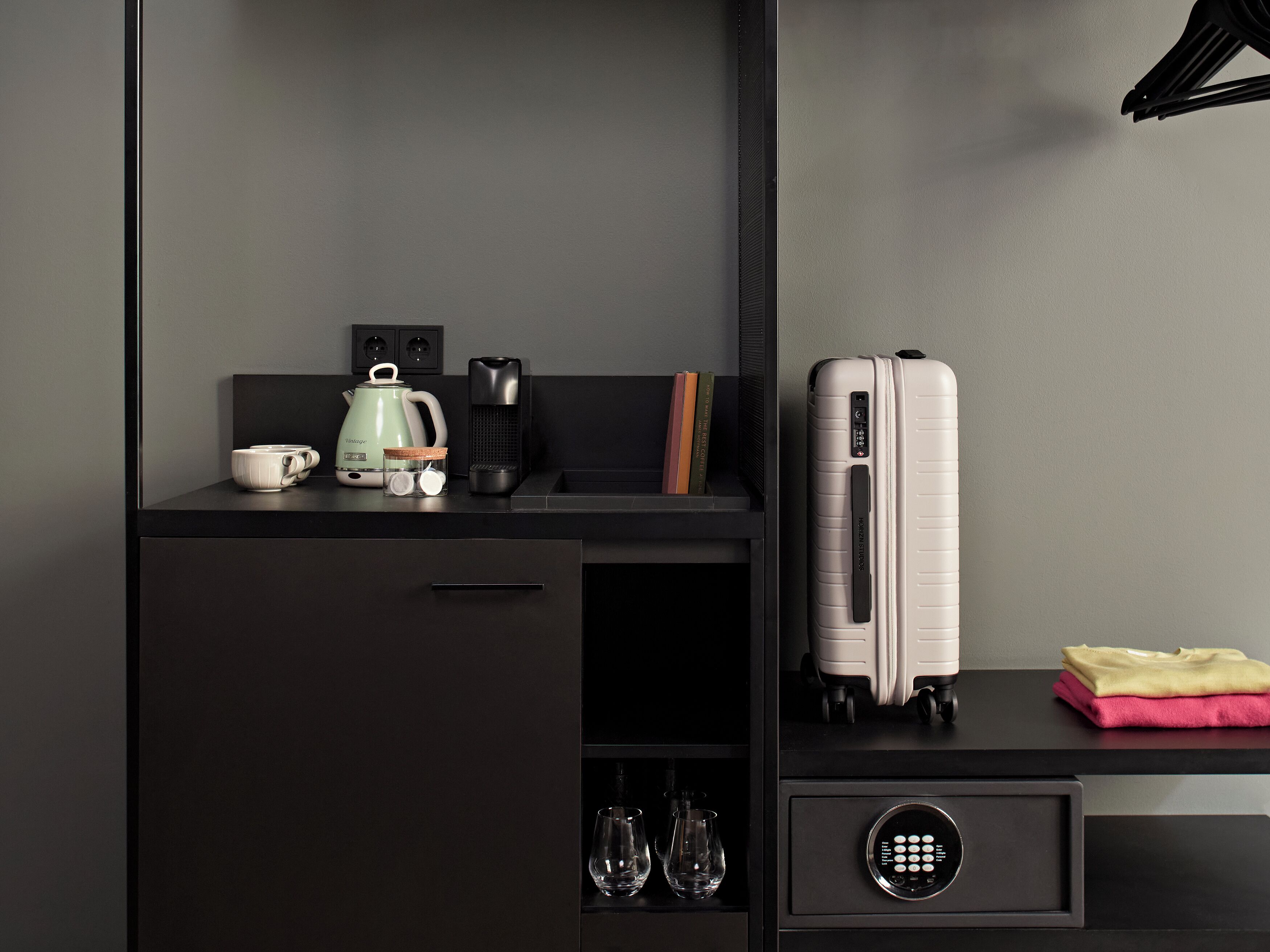 Una moderna camera d'albergo con una scrivania scura, un bollitore, un telecomando, una valigia e una cassaforte.