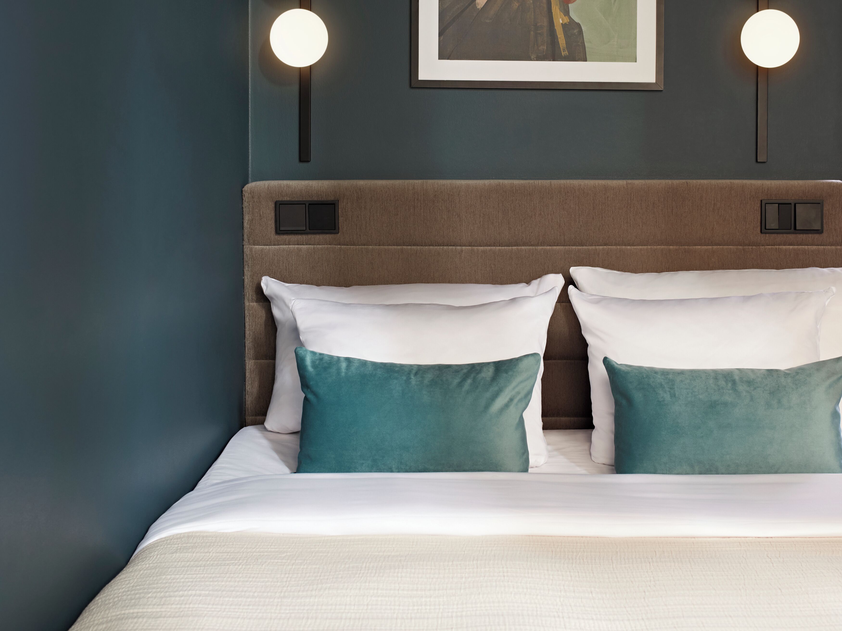 Una cama bien hecha con almohadas blancas y azul verdosas, un cabecero marrón y dos lámparas fijadas a la pared.