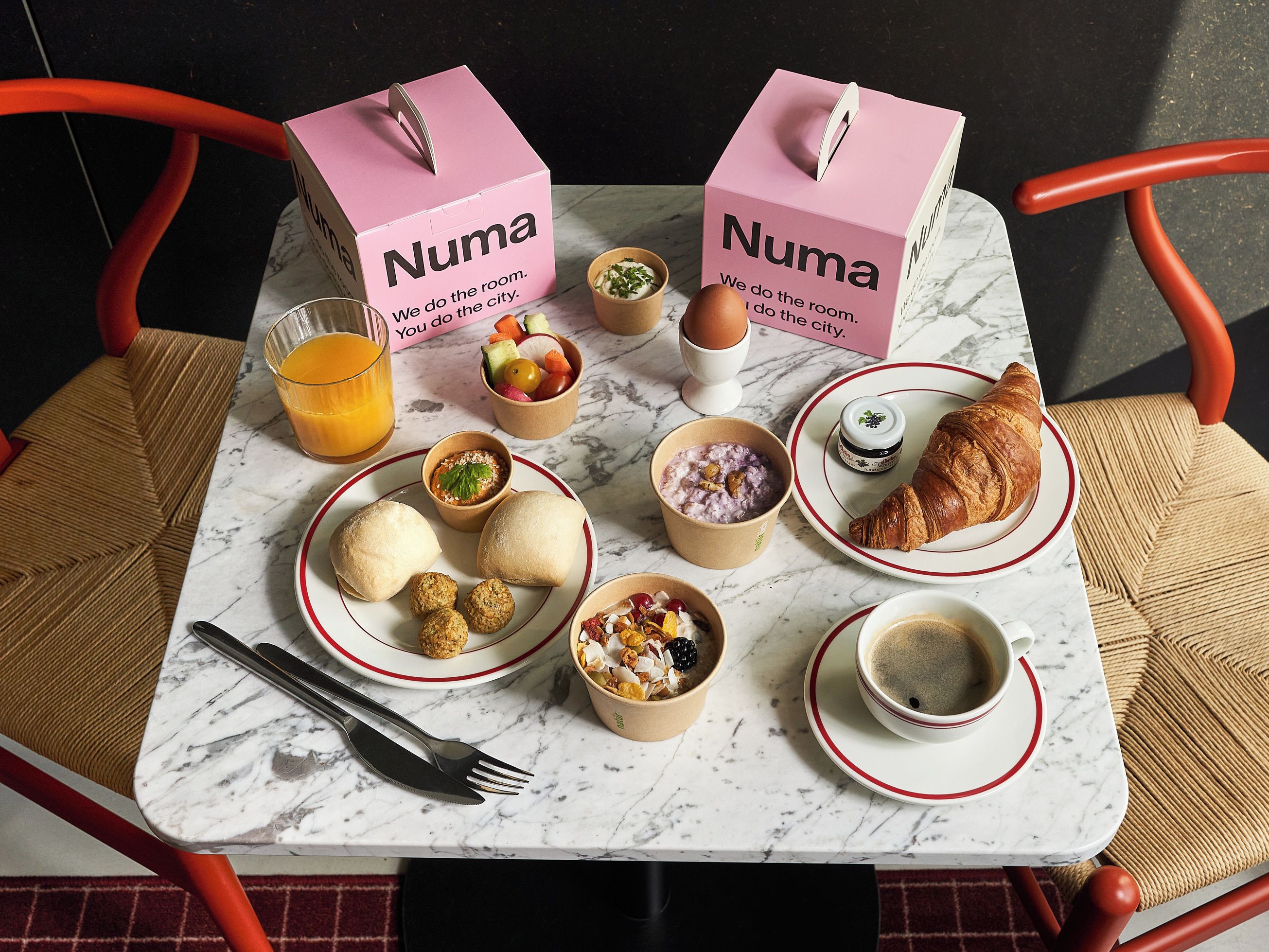 Une table de marbre dressée pour le petit-déjeuner, avec deux boîtes roses étiquetées « Numa », un croissant et divers autres éléments pour le petit-déjeuner.