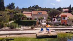 A waterfront property with clear water, a dock with lounge chairs, and a stone pathway leading to the buildings.