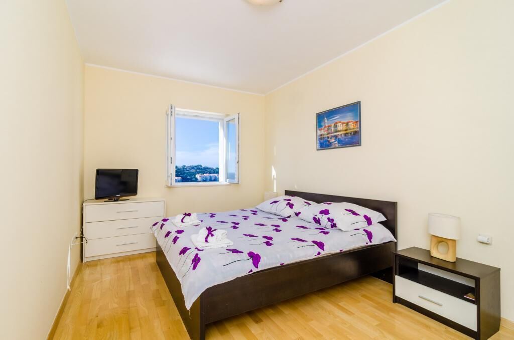 A bedroom with a bed, a TV on a dresser, a window with a view, and a painting on the wall.