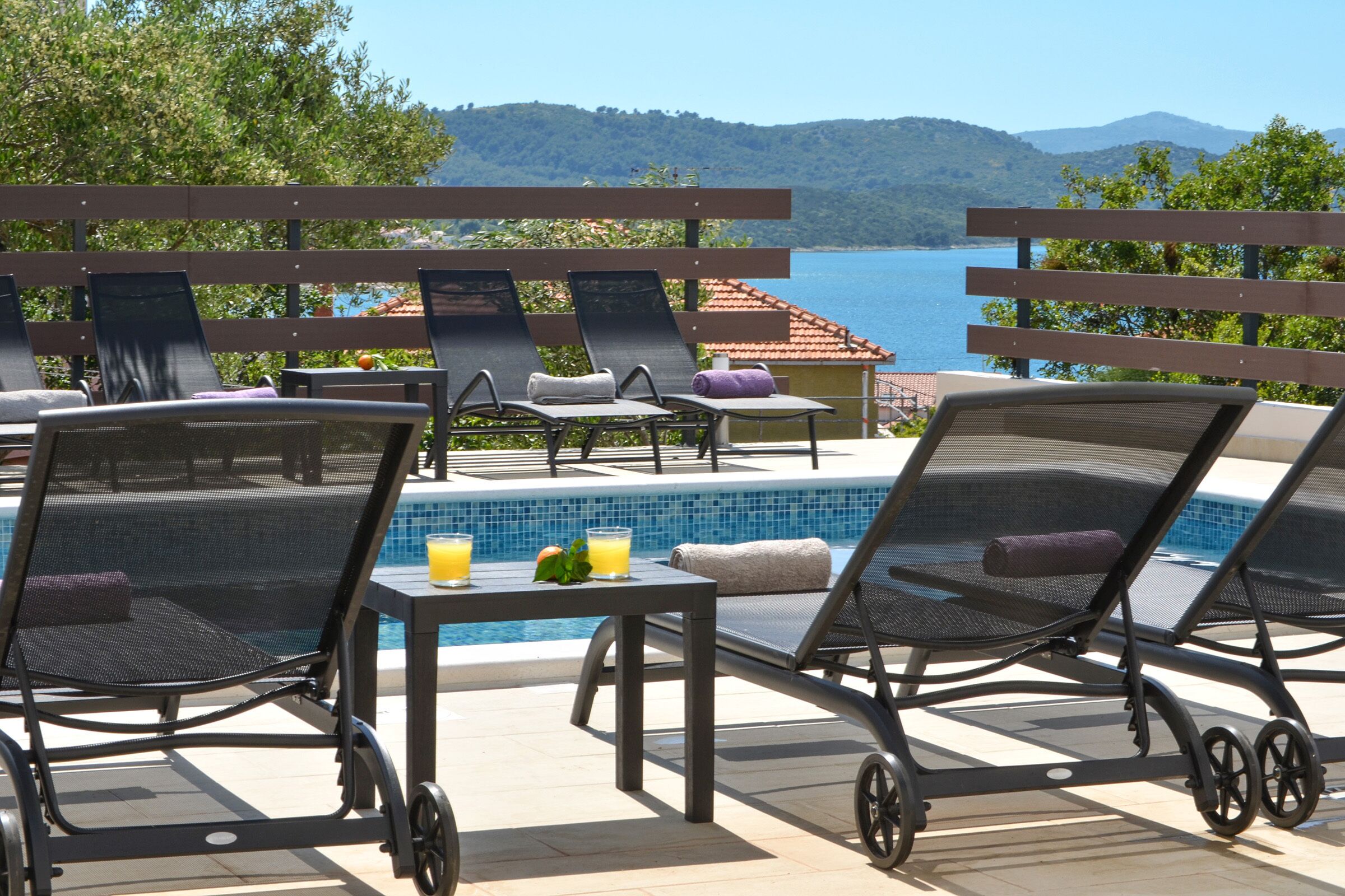 A poolside area with lounge chairs and a table, overlooking a body of water and hills.