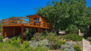 A wooden deck structure with a covered seating area, surrounded by lush greenery and flowering plants.
