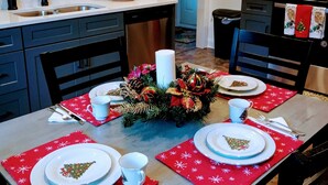 Un'area pranzo con tavolo imbandito per quattro persone, con sottopiatti rossi decorati con fiocchi di neve, piatti bianchi con motivo natalizio e un centrotavola con verde e decorazioni.