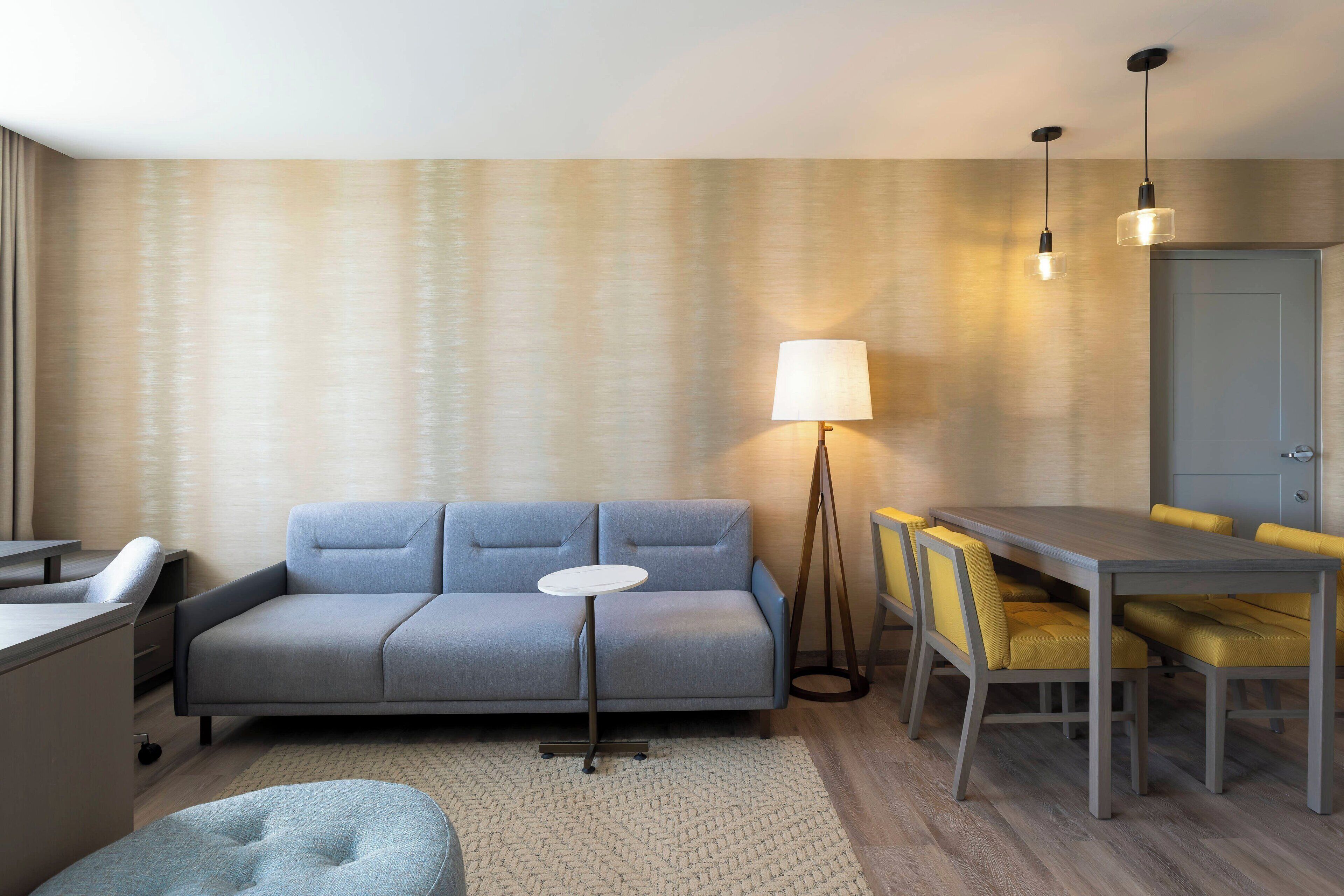 A modern hotel room with a grey sofa, a small round table, a dining table with yellow chairs, and a floor lamp.