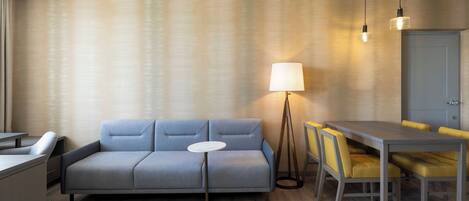 Une chambre d’hôtel moderne avec un canapé gris, une petite table ronde, une table à manger avec des chaises jaunes et un lampadaire.