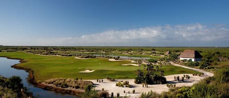 A golf course with a pond, a thatched-roof building, and a wooden bridge.