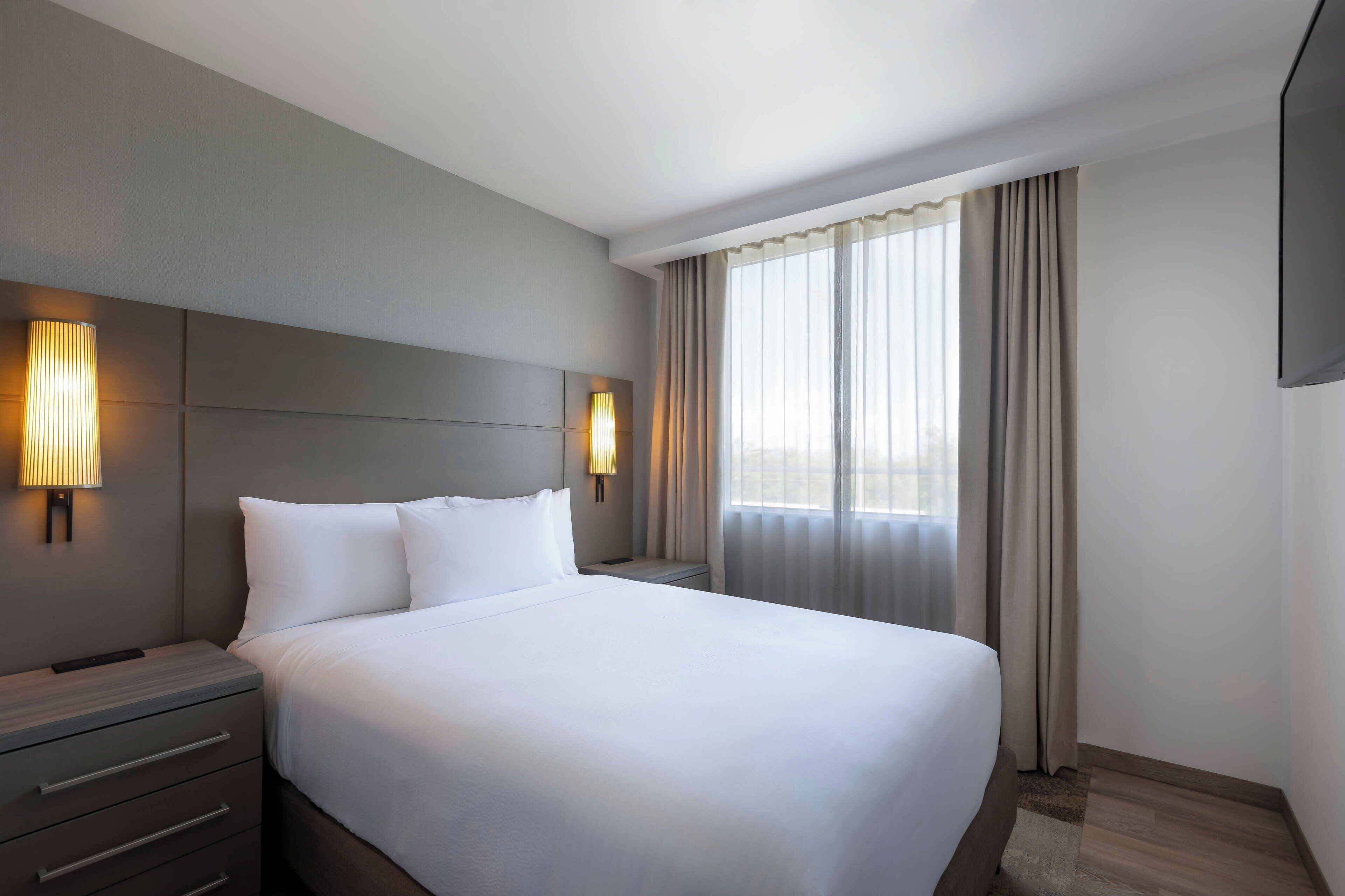 A hotel room with a large bed, two bedside tables, a TV mounted on the wall, and a window with curtains.