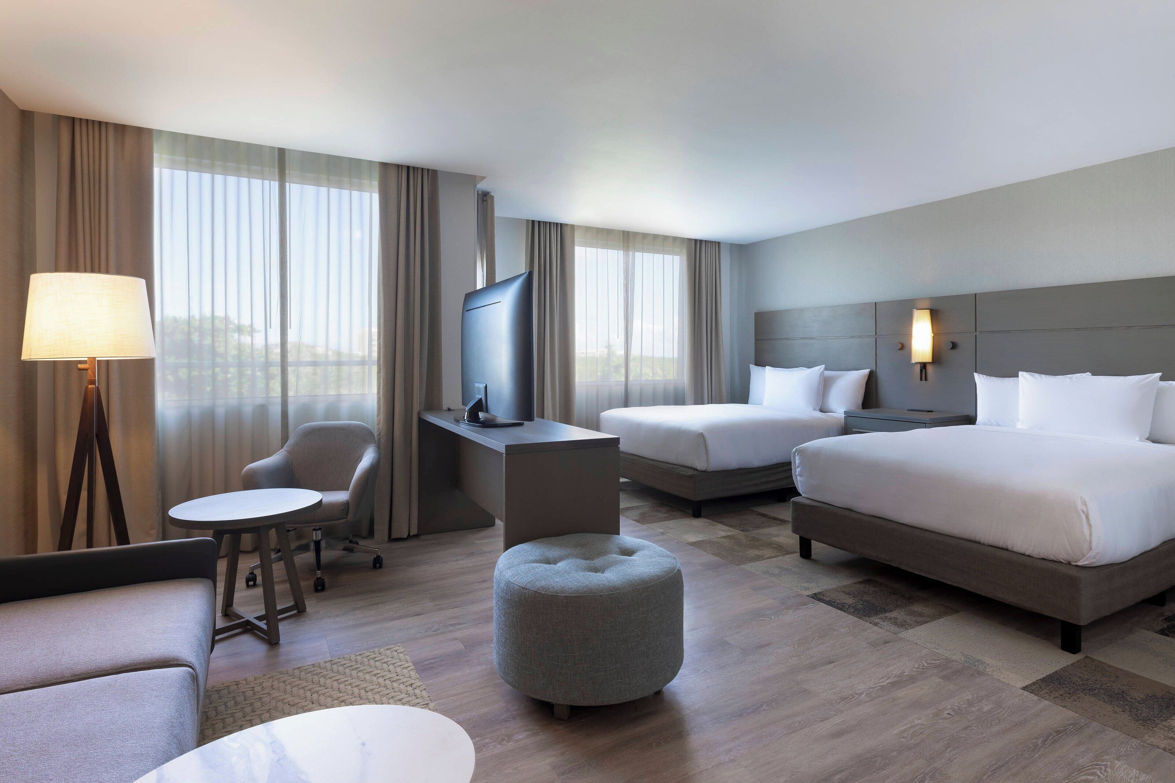 A hotel room with a bed, two bedside tables, a desk with a chair, a round ottoman, a sofa, and a TV.