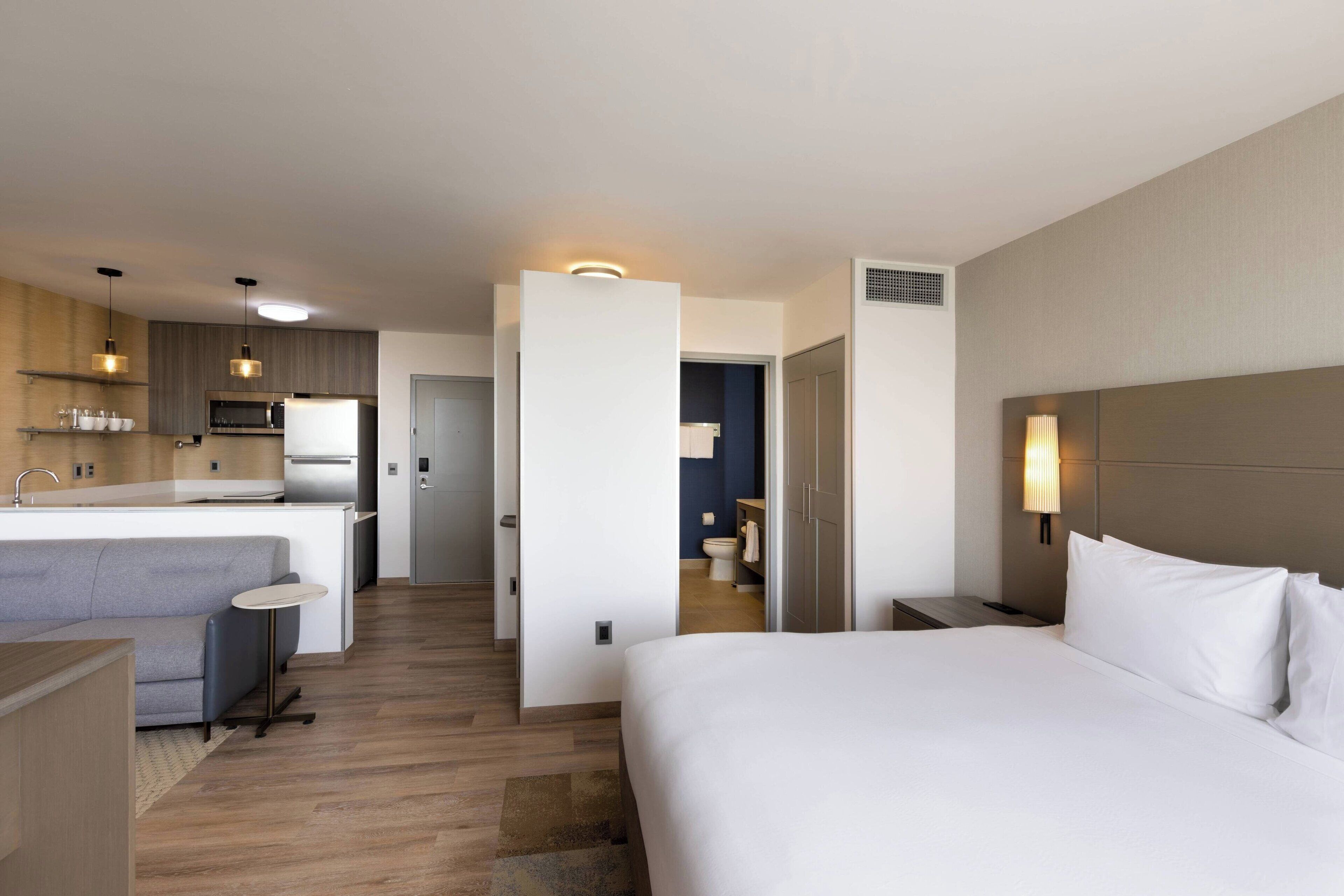 Una camera d'albergo moderna con un letto grande, un piccolo angolo salotto, una kitchenette e un bagno visibile attraverso una porta aperta.