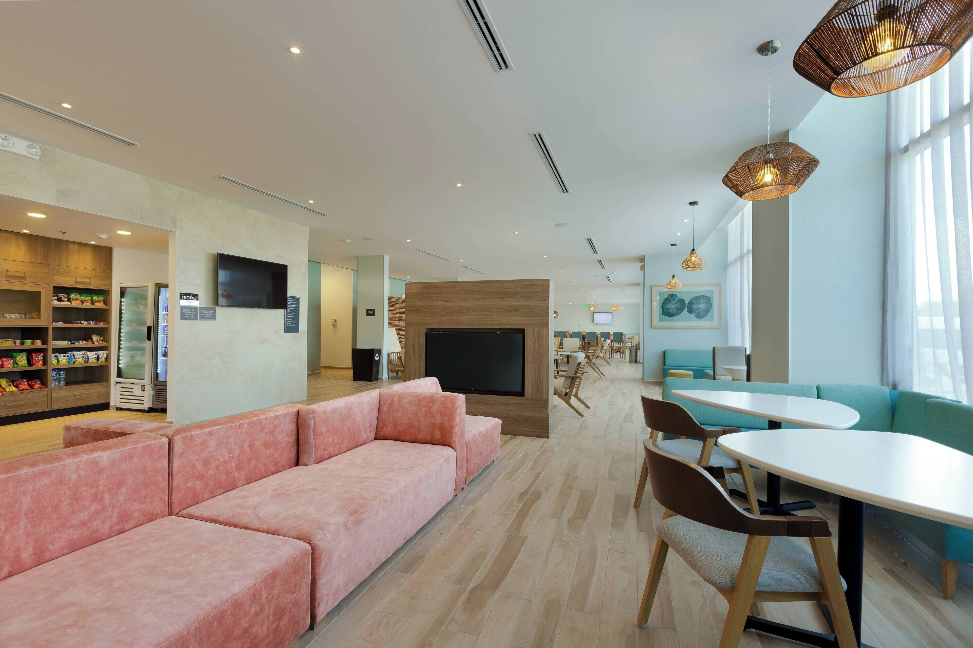 A modern hotel lobby with a pink sofa, a TV, and a dining area with tables and chairs.