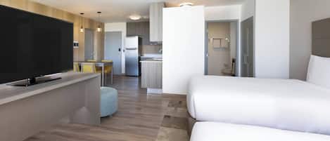 A modern hotel room with a flat-screen TV, a dining area, a kitchenette, and a separate bathroom.