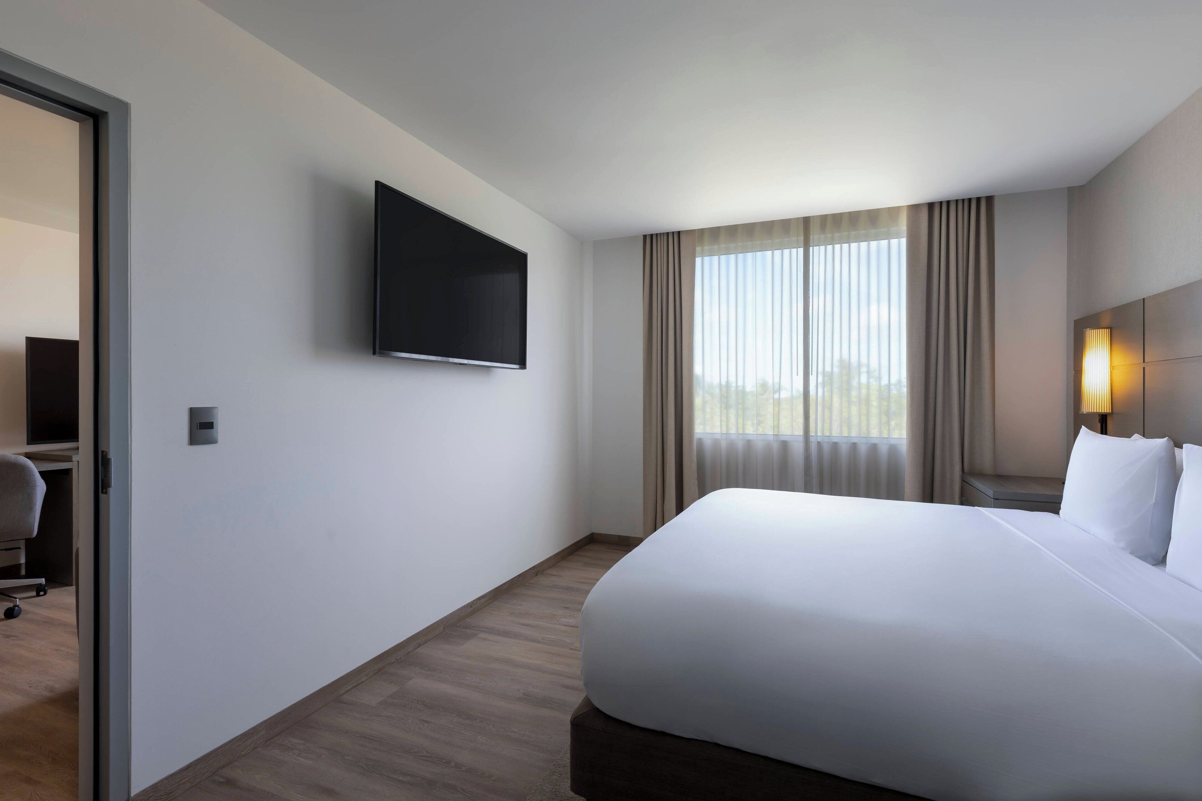Habitación de hotel con una cama grande, un televisor de pantalla plana montado en la pared, un escritorio con una silla y una ventana con cortinas.