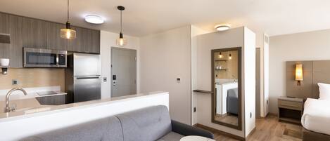 A modern hotel room with a kitchenette, a grey sofa, a small round table, a bed, and a bathroom with a mirror and sink.