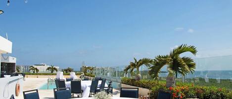 A rooftop dining area with tables and chairs, a pool, and a view of the ocean.