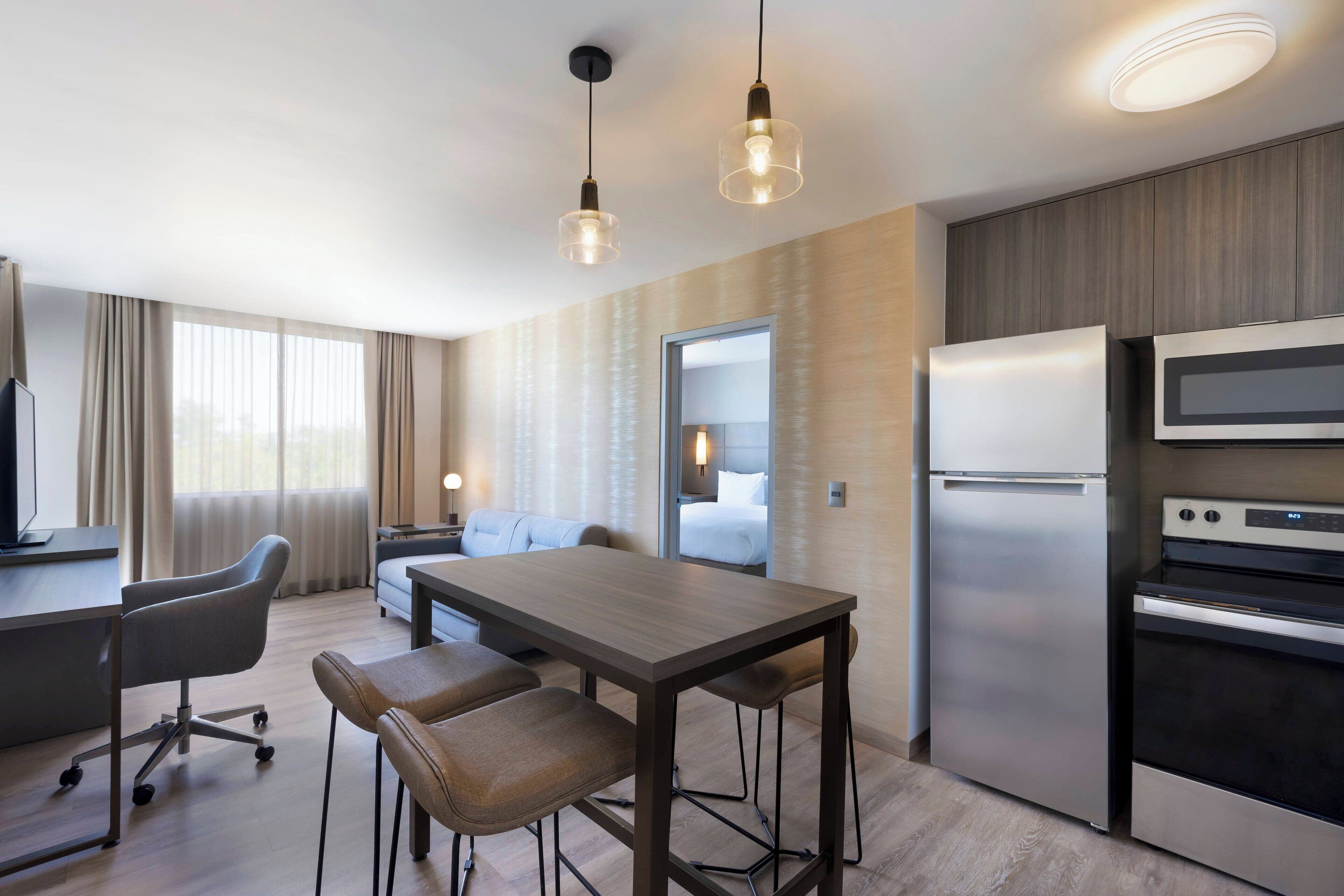 A modern hotel room with a wooden dining table, two chairs, a sofa, a TV, a refrigerator, and a microwave.
