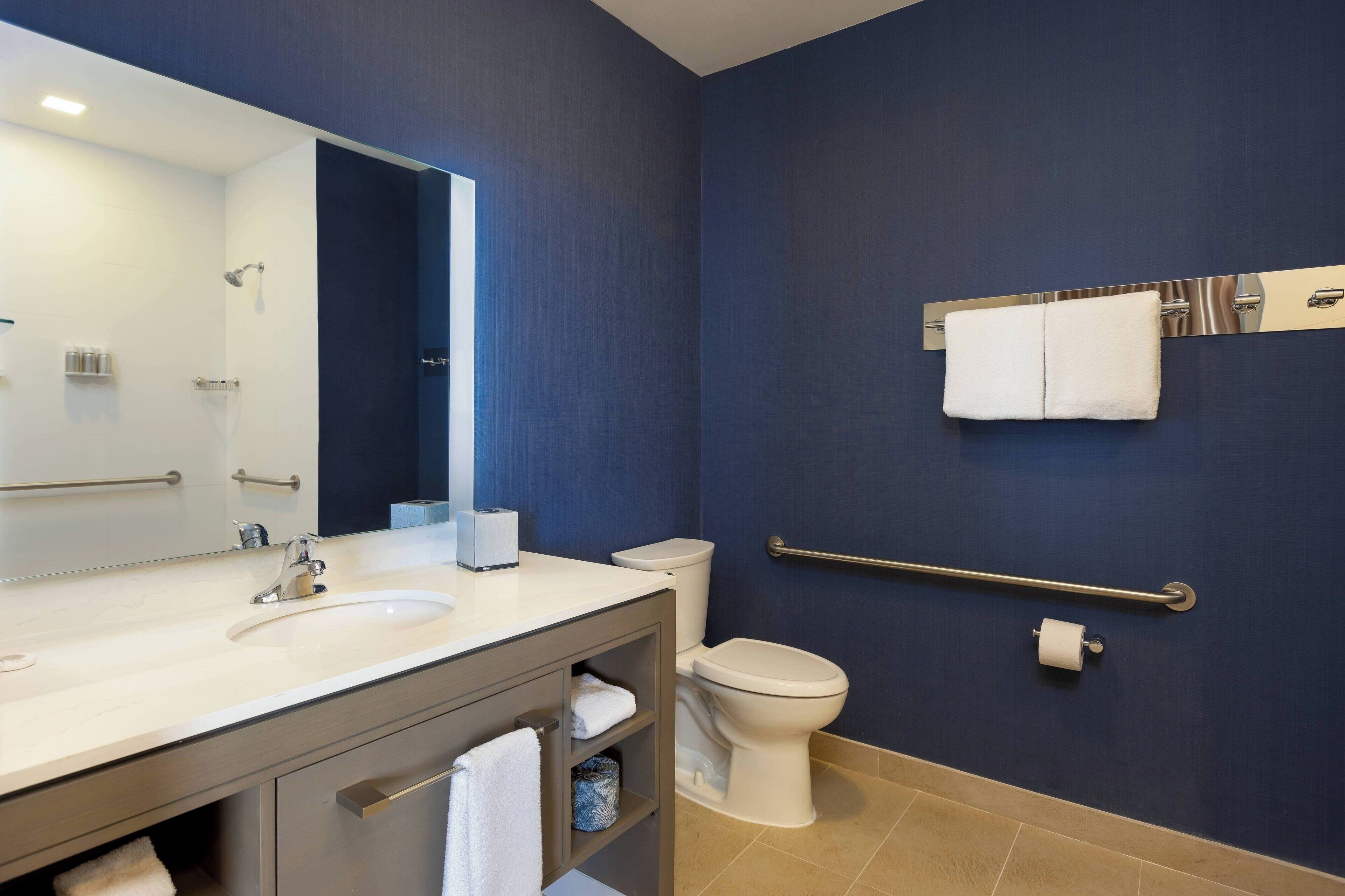 A modern bathroom with a large mirror, sink, and toilet. There is a towel rack on the wall and a grab bar next to the toilet.