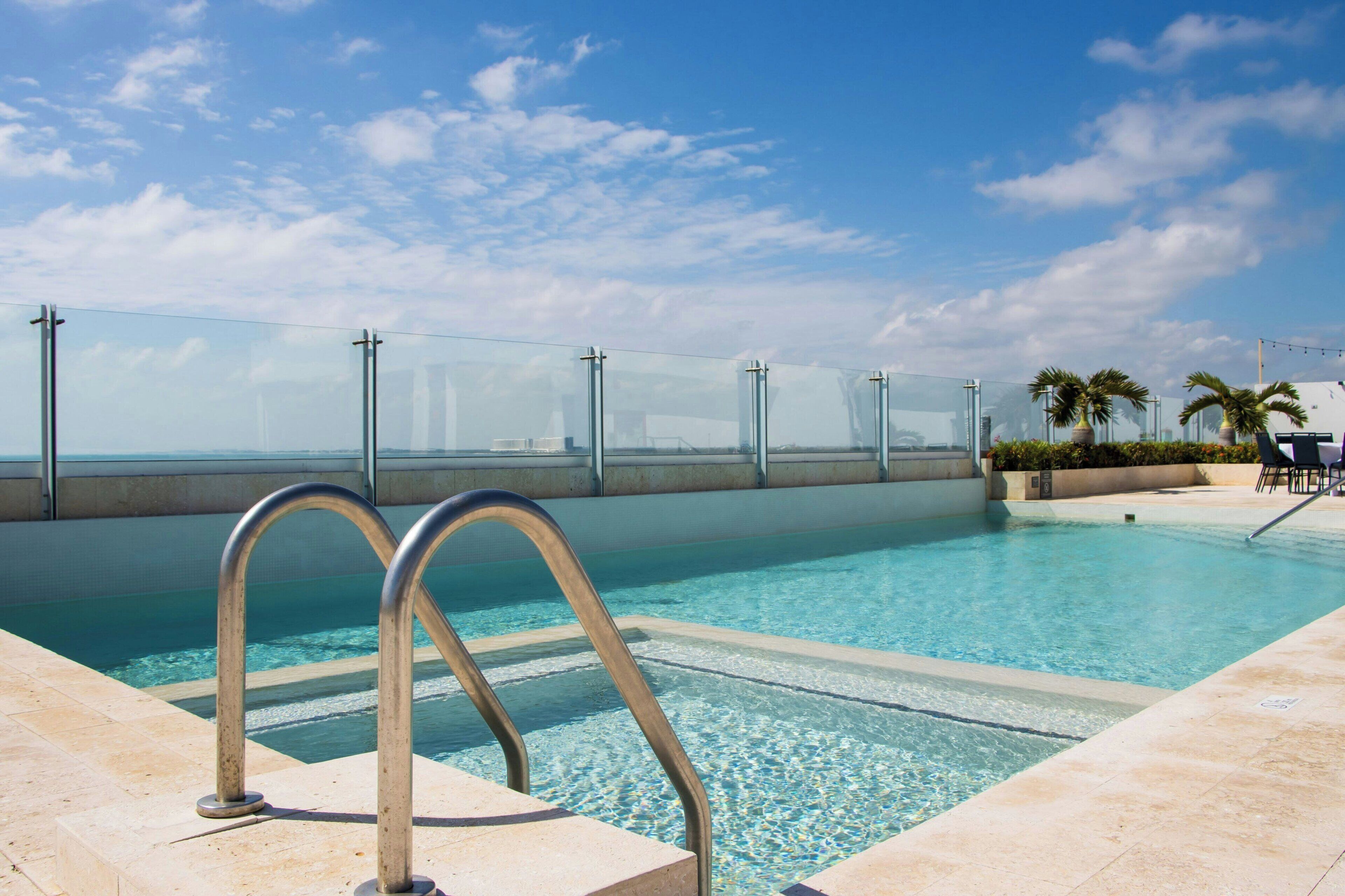 A clear swimming pool with a glass barrier, palm trees, and a view of the city skyline.