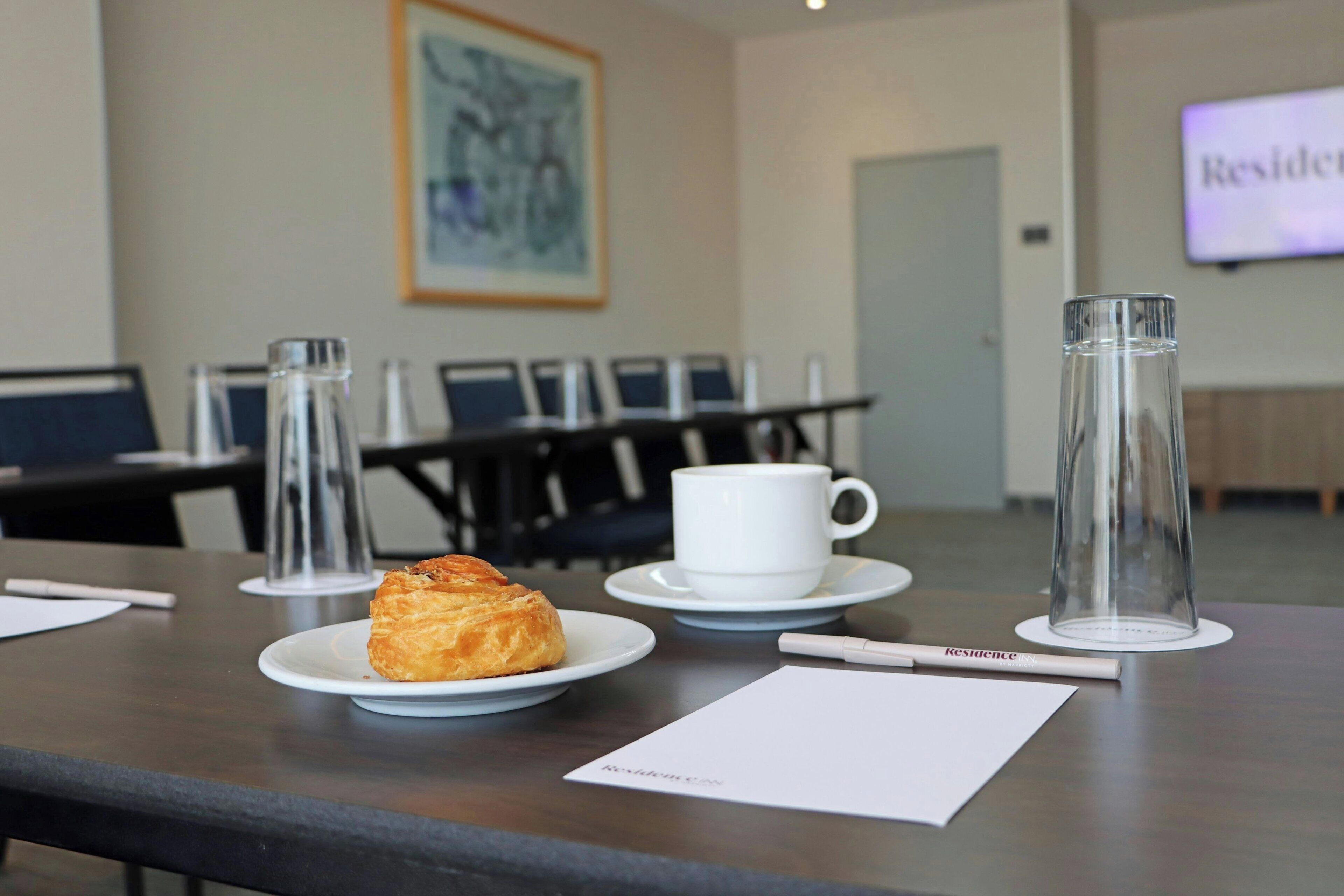A conference room with a table set for a meeting, including a plate with a pastry, a cup, and a glass.