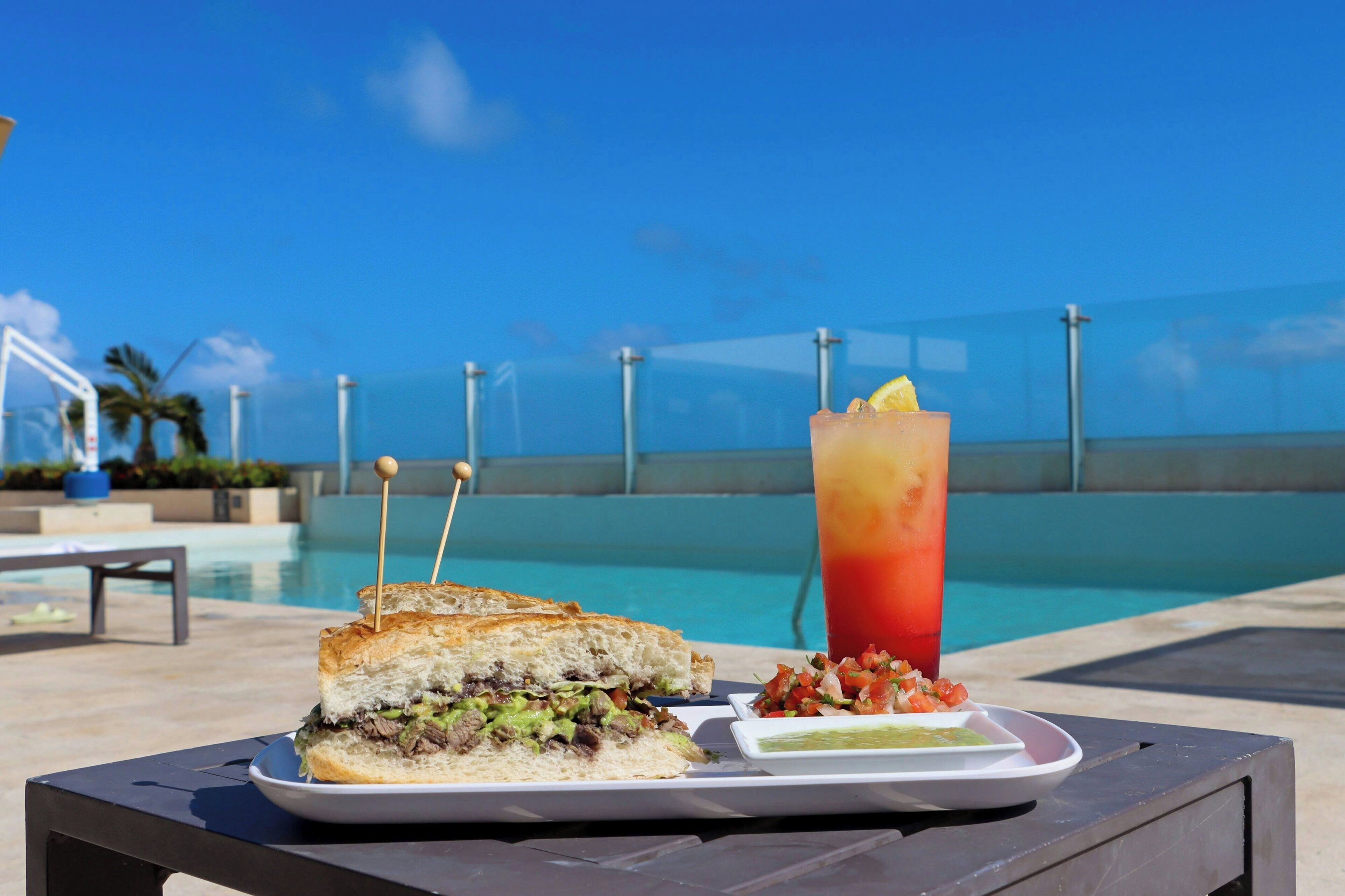 Una mesa con un sándwich, una bebida y un recipiente pequeño de salsa junto a una piscina.