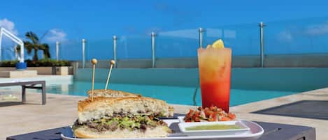 Una mesa con un sándwich, una bebida y un recipiente pequeño de salsa junto a una piscina.