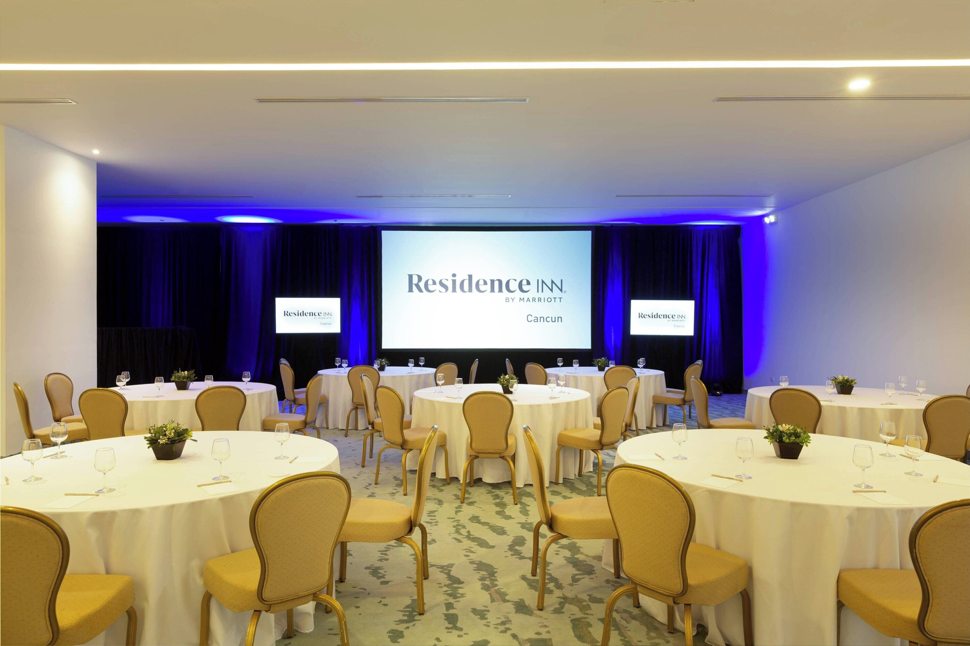 Sala de conferências com várias mesas redondas, cadeiras e uma tela grande exibindo 'Residence IN Cancun'.
