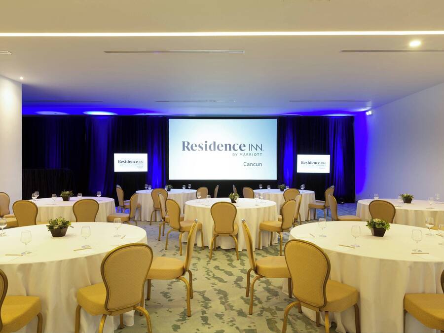 Sala de conferências com várias mesas redondas, cadeiras e uma tela grande exibindo 'Residence IN Cancun'.