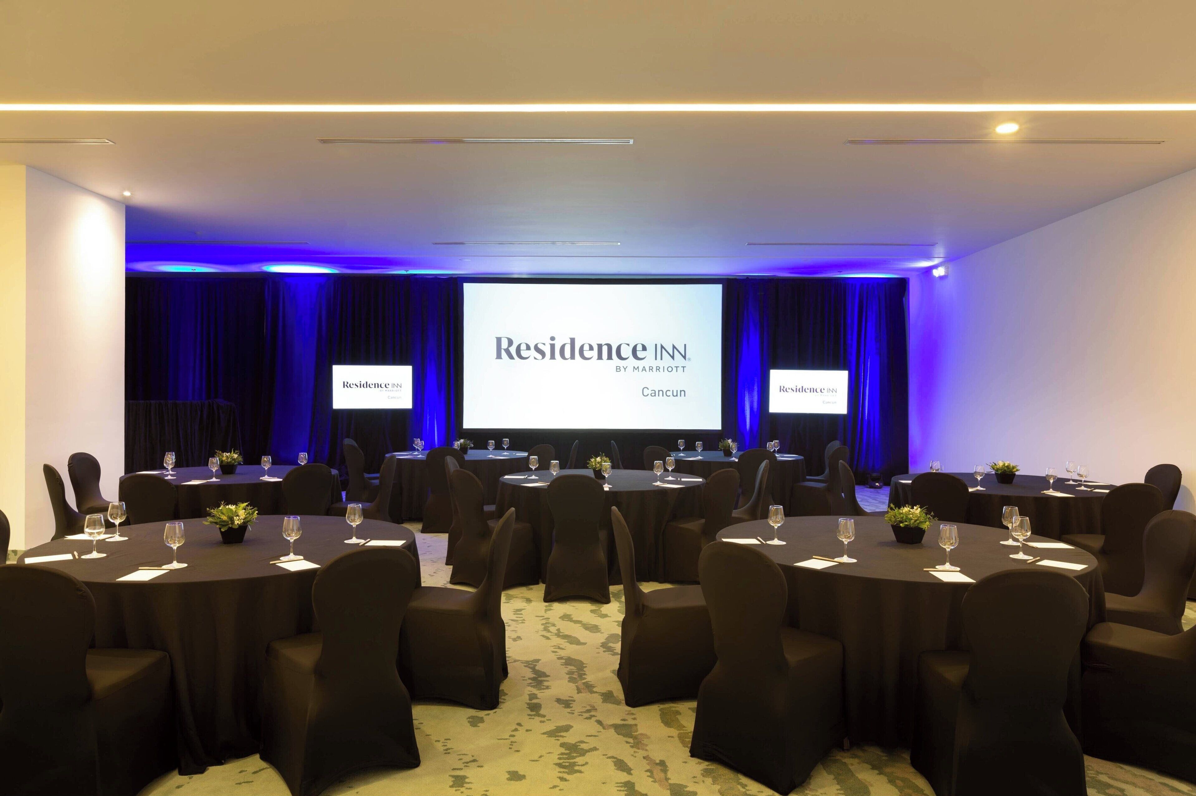 Un salón de conferencias con una pantalla grande mostrando 'Residence IN by Marriott Cancun' y varias mesas dispuestas para una reunión.