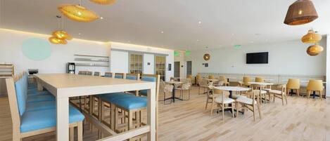 Un grand espace à manger avec des tables en bois et des chaises rembourrées bleues, agrémenté de lampes en osier suspendues et d’un téléviseur à écran plat fixé au mur.