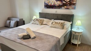 En pænt redt seng med en velkomsthilsen, to puder med broderet tekst 'HOME', en grå sofa, et lille natbord med lampe, og et farverigt abstrakt billede på væggen.