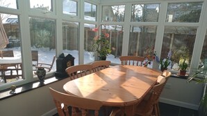 Ein Wintergarten mit Holztisch und -stühlen, großen Fenstern und Blumentöpfen.
