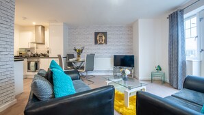 Una sala de estar moderna con sofá, una mesa de centro y una televisión. Se observa una cocina al fondo, con estufa, horno y armarios.