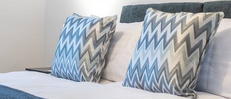 Una cama bien hecha con almohadones estampados y una pared lisa con dos cuadros enmarcados.