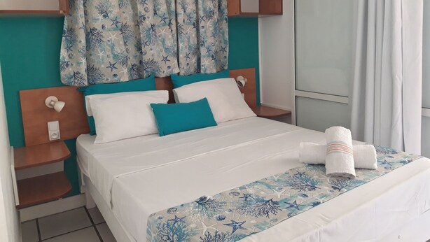 Cama bem arrumada com colcha branca e azul estampada, duas almofadas brancas e uma almofada de destaque azul. Há duas mesas de cabeceira com luminárias, uma pequena prateleira sobre a cama e uma janela com cortinas.
