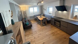 A modern kitchen with a built-in stove, a dining area with a wooden table and chairs, and a seating area with a sofa and cushions. - Description (Schwendau)
