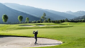A golf course with a player in a sand bunker, surrounded by lush greenery and mountains. - Description (Schwendau)